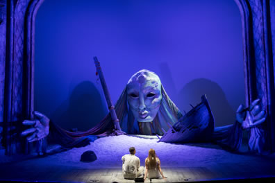The giant head of Juno is at the back of the stage, with giant hands spread out to the side; the head appearing between the shipwrecked bow and mast on a mound of sand, with the two lovers, backs to us, in the foreground. The whole photo has a blue tone.